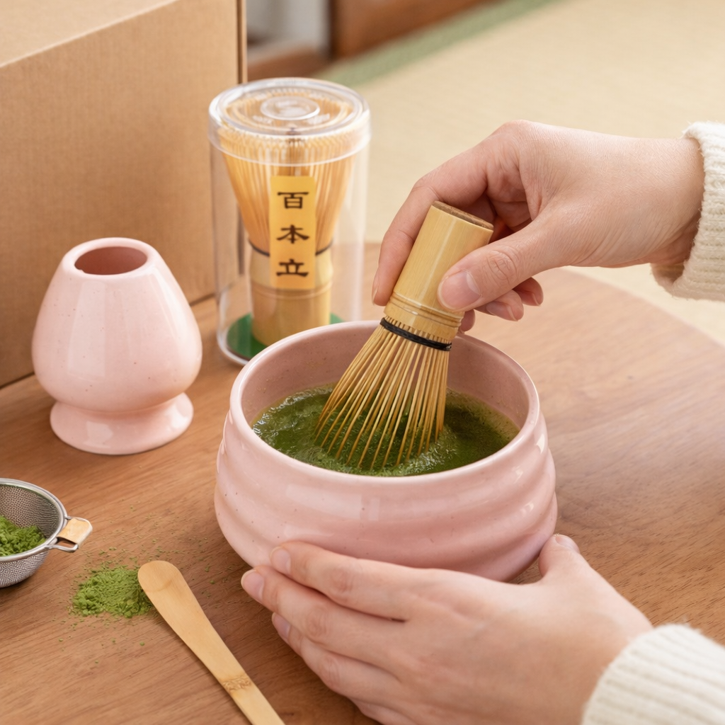A person whisks green matcha in a pink bowl using a bamboo whisk, with matcha powder and a wooden spoon from the Różowy zestaw matcha 4 elementy displayed on the wooden table.