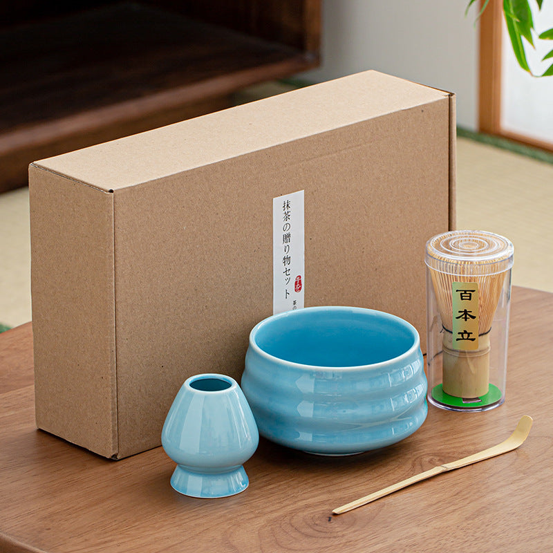 The Niebieski zestaw matcha 4 elementy displayed on a wooden table includes essentials: a blue tea bowl, matching blue whisk holder, bambusowa miotełka in a clear container, and a bamboo tea scoop.