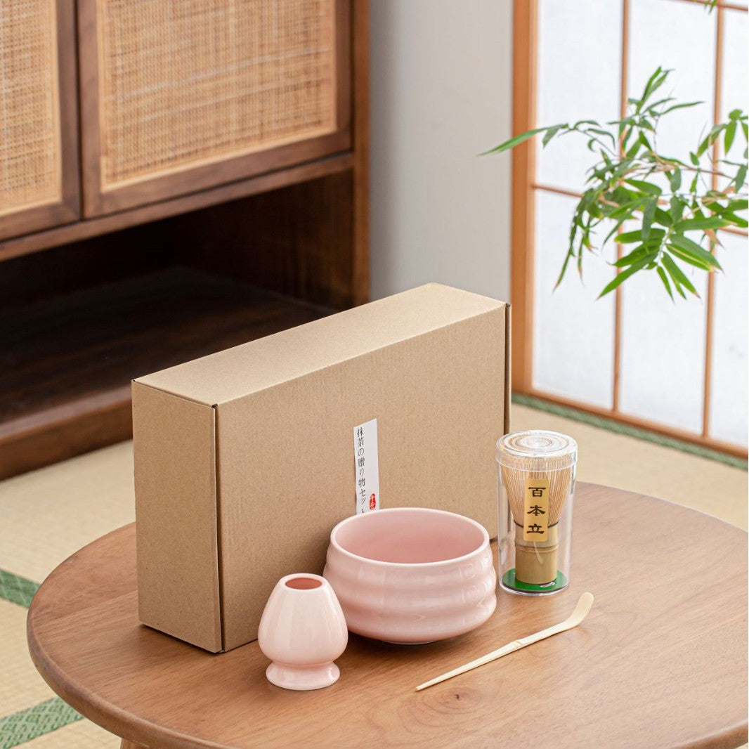 The Różowy zestaw matcha 4 elementy is displayed on a wooden table with a pink tea bowl, bamboo whisk in a container, pink whisk holder, bamboo scoop, and brown gift box—Japońska herbata magic shown with plant and shoji screen in the background.