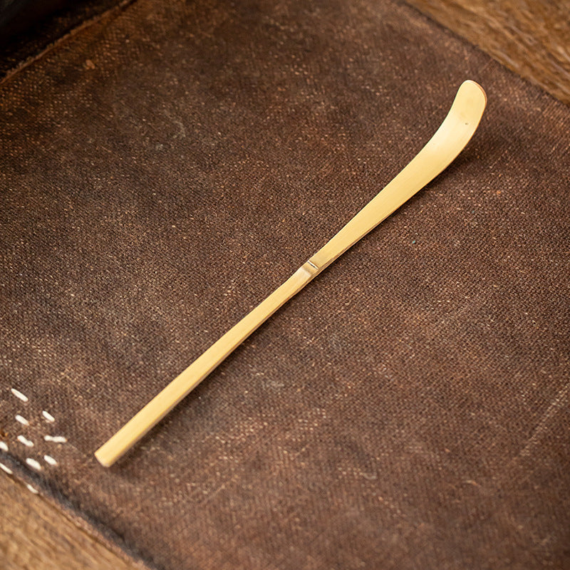 A bamboo tea scoop with a curved end rests on textured brown fabric—a great tool for preparing matcha with your Zielony zestaw matcha 4 elementy.