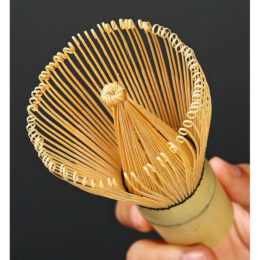 A hand holding a traditional chasen from the Zielony zestaw matcha 4 elementy set prepares matcha tea, with the intricate bamboo prongs highlighted against a dark background.