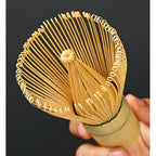 A hand holds a bambusowa miotełka (chasen) with delicate, curved tines, set against a black background—highlighting the elegance of preparing matcha with the Biało-różowy zestaw matcha 4 elementy.