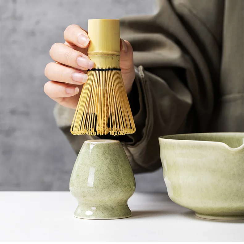 A person holds a bambusowa miotełka above a green ceramic holder, with a matching bowl nearby—elements from the Zielony zestaw matcha 4 elementy, perfect for your matcha ceremony, all arranged on a white surface.