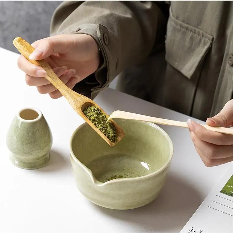 A person in a green coat uses a bamboo scoop to add matcha from the Zielony zestaw matcha 4 elementy into a ceramic bowl, with a bambusowa miotełka and small ceramic container nearby on a white surface.
