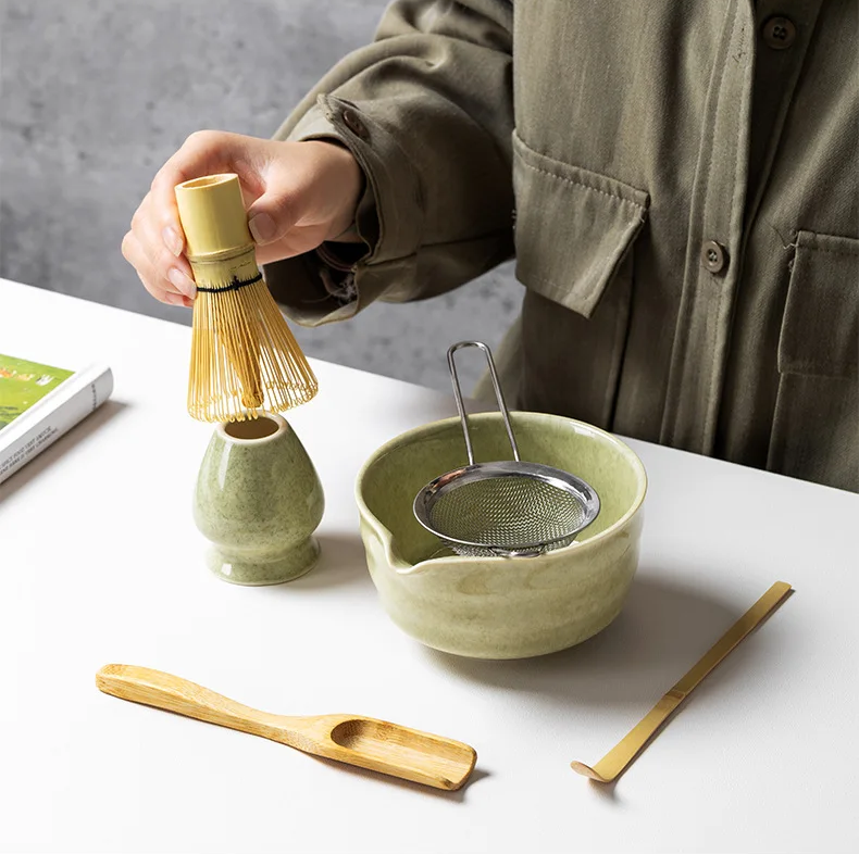 A person holds a bambusowa miotełka over a green ceramic holder, with the Zielony zestaw matcha 4 elementy—featuring a green bowl, metal strainer, and wooden tea utensils—arranged on a white table.